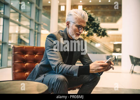 Mature businessman sitting in office lounge and using mobile phone. Senior executive texting on phone in office lobby. Stock Photo