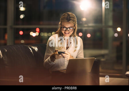 Happy young woman sitting in office and reading text message on her smart phone. businesswoman using phone while working late in office. Stock Photo