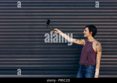 Female blogger making a video using her smart phone mounted on flexible tripod. Young woman vlogging on background of a store shutter. Stock Photo