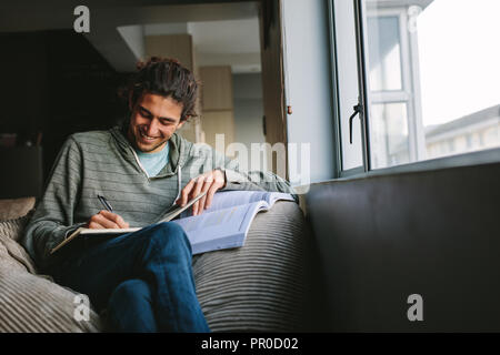 Smiling man sitting comfortably at home writing in a book. Student writing notes from a text book sitting on couch beside a window. Stock Photo