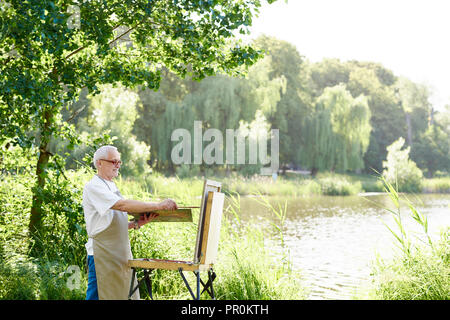 Serious male artist holding brush and painting picture on easel on open air. Senior painter, wearing in apron and modern glasses, using palette with paints working on his new masterpiece against lake. Stock Photo