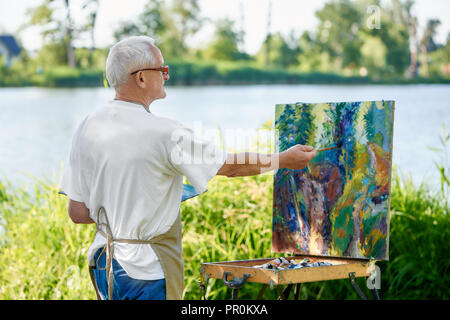 Rear view of creative painter paints colorful abstract painting on open air. Senior painter, wearing in apron and modern glasses, using palette with paints working on his new masterpiece against lake. Stock Photo