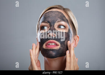 Close-up Of A Happy Woman Using Activated Charcoal Face Mask On Grey Background Stock Photo