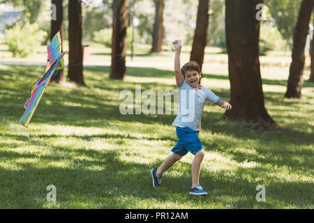 adorable happy boy holding colorful kite and running on grass in park Stock Photo