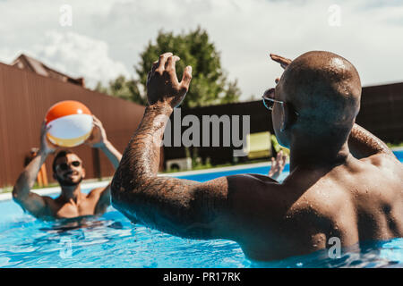 multicultural men playing with beach ball in swimming pool Stock Photo