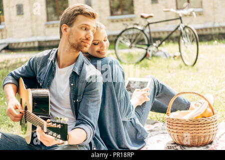 handsome young man playing guitar for his smiling girlfriend while having picnic at park Stock Photo