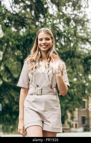 smiling young woman in stylish overall looking at camera and waving with hand Stock Photo