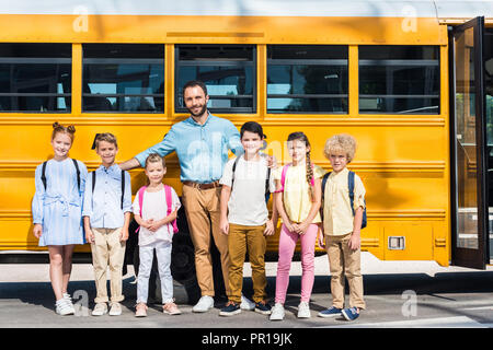 schoolchildren and teacher standing together in front of school bus and looking at camera Stock Photo