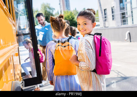 little schoolgirl entering school bus with classmates while teacher standing near door Stock Photo