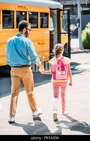 rear view of father and daughter with backpack holding hands and walking to school bus Stock Photo
