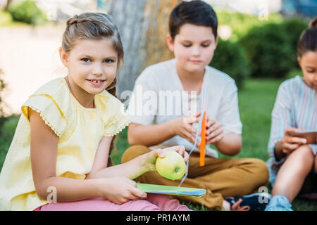 adorable little schoolgirl with apple and book sitting on grass with classmates Stock Photo