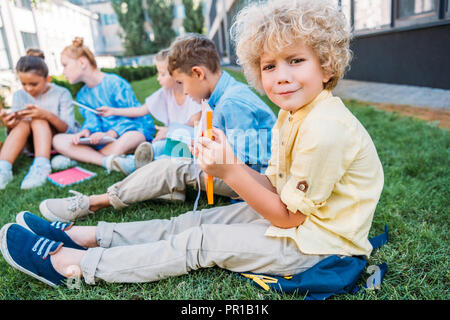 confused curly schoolboy holding book while sitting on grass with classmates Stock Photo