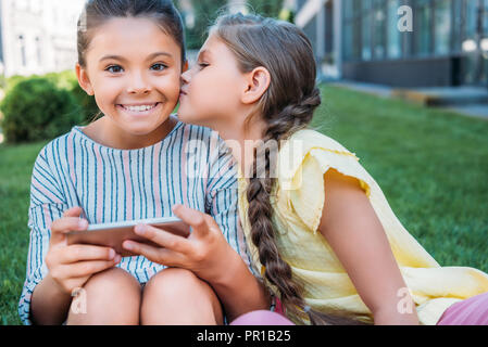 adorable little schoolgirl with smartphone looking at camera while her friend kissing her cheek Stock Photo
