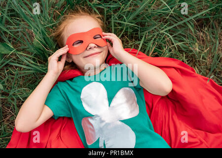 overhead view of kid in red superhero cape and mask lying on green grass Stock Photo