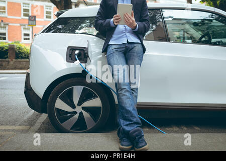 Businessman using digital tablet while charging electric car at charging station Stock Photo