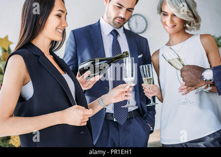 handsome businessman pouring champagne to multicultural colleagues glasses in office Stock Photo