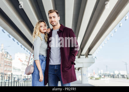 stylish couple in autumn outfit hugging under bridge in city and looking at camera Stock Photo