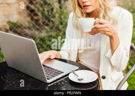 cropped image of woman using laptop and holding cup of coffee at table in garden Stock Photo
