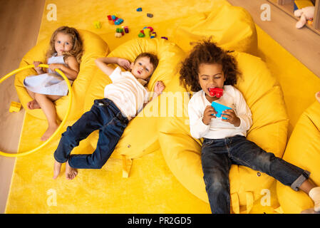 high angle view of multiethnic kids lying on bean bag chairs with toys in kindergarten Stock Photo