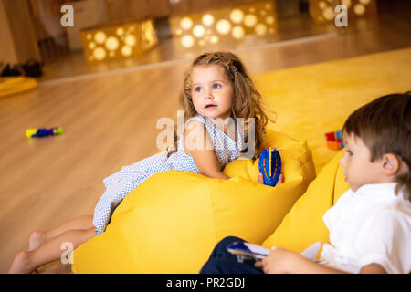adorable children with toys lying on yellow bean bag chairs in kindergarten and looking away Stock Photo