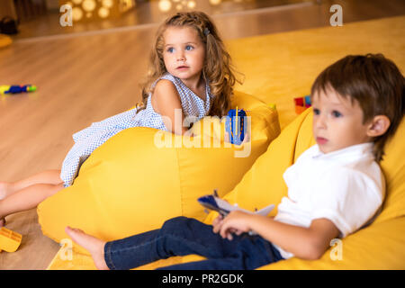 adorable kids with toys lying on yellow bean bag chairs in kindergarten and looking away Stock Photo
