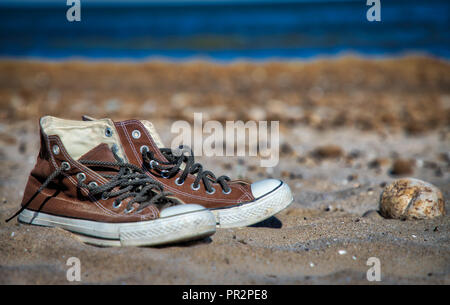 A pair of old worn brown canvas style retro basketball sneakers (one shoe in focus, the other in soft focus) on the sand at the beach with the water i Stock Photo