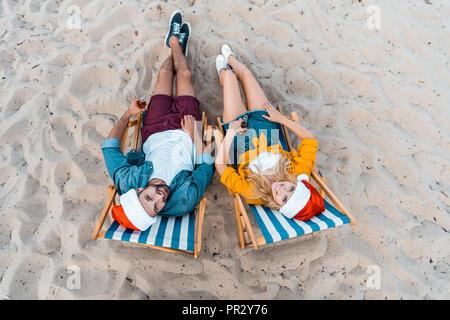 high angle view of couple in santa hats lying on sun loungers with beer in bottles on sandy beach Stock Photo