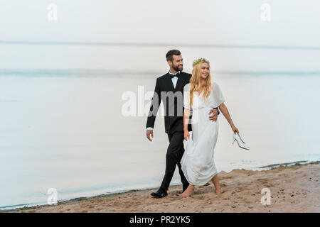 wedding couple hugging, walking on beach and looking away, bride holding high heels in hand Stock Photo