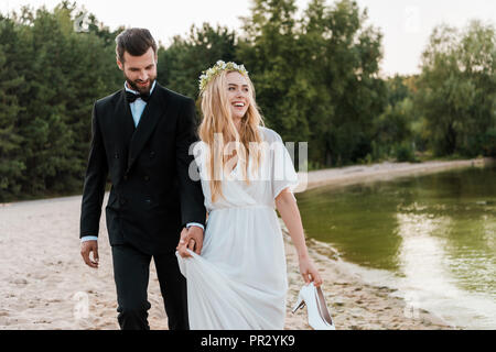 wedding couple holding hands and walking on beach, laughing bride holding high heels in hand Stock Photo