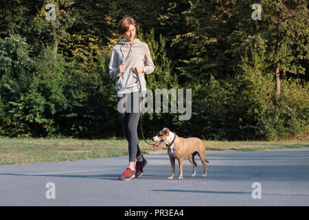 Woman trains her dog to run along during jogging. Young fit female and staffordshire terrier dog at a morning walk doing physical excercise