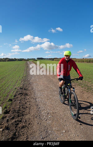 Closeup of smiling Male Cyclist wearing red and a safety helmet cycling towards the camera along The Pennine Way, Wortley, South Yorkshire, England Stock Photo