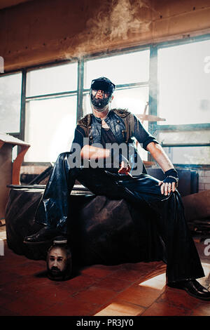 The man in a leather cloak and trousers with ornamentals on his head smoking a cigarette near the jar with a head of a man indoors. Brutal dark style Stock Photo