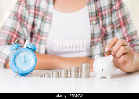 Woman hand putting money coin into piggy bank and stack of coins and alarm clock. Saving money wealth and financial concept. Stock Photo