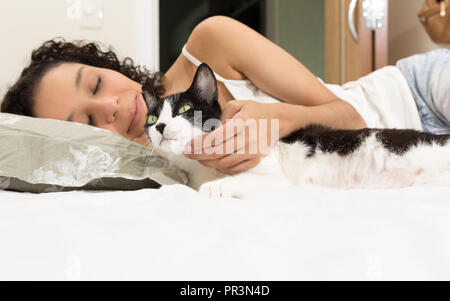 Portrait of mixed race woman lying in bed and hand petting black and white cat. Room interior on background. Concept of love to animals, pets, lifesty Stock Photo