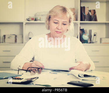 Mature woman sits at the table and looks through bills and checks Stock Photo