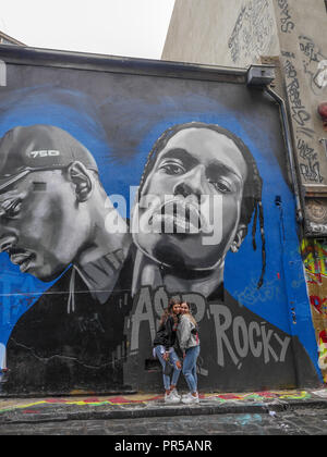 Vertical shot of two teenage girls posing in front of a large graffiti portrait of two African American men painted on a wall in Hosier Lane. Stock Photo
