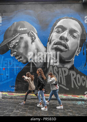 Vertical shot of three teenage girls walking in front of a beautiful portrait of two young black men painted on wall in Hosier Lane in Melbourne. Stock Photo