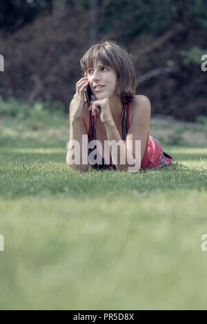 Retro effect faded and toned image of a young woman chatting on a mobile phone as she lies on her stomach on lush green grass, low angle view. Stock Photo