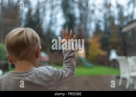 Young boy staring out of a view window into a garden with his hand on the glass in a rear view with copy space. Stock Photo