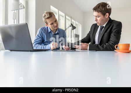 Two businessmen discussing data on a tablet as they sit enjoying coffee at a table in the office in a low angle view with copy space. Stock Photo