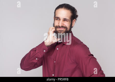 Portrait of confused handsome man with dark collected long hair and beard in red shirt standing and touching his face, thinking and looking away. indo Stock Photo