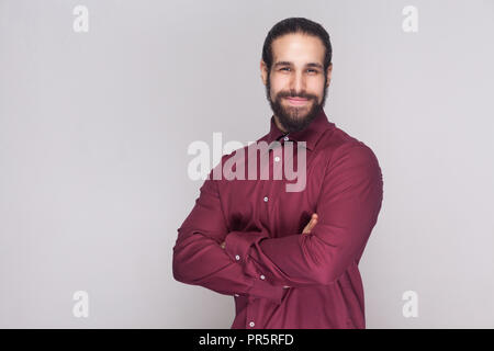 Portrait of handsome businessman with dark hair and beard in red shirt standing with crossed hands, looking at camera with satisfied face and smiling. Stock Photo