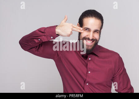 Portrait of crazy handsome man with dark collected long hair and beard in red shirt standing and holding hand on head with gun gesture. indoor studio  Stock Photo