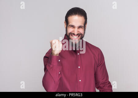 Yes, i win. Portrait of handsome man with dark hair and beard in red shirt standing and looking at camera with winner face and celebrating victory. in Stock Photo