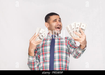 Happy joyful handsome young adult businessman in colorful checkered shirt with blue tie standing, holding fan of dollars and winking with open mouth.  Stock Photo