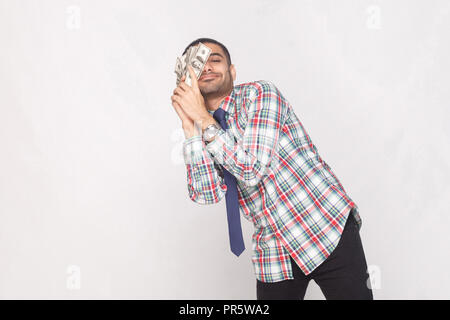 Dream come true! Satisfied rich handsome bearded businessman in colorful checkered shirt with blue tie standing and lean against to cheek fan of money Stock Photo