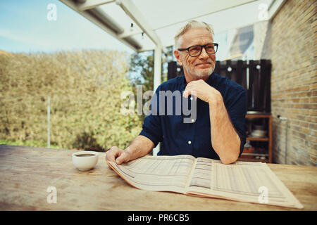 Smiling senior man drinking a cup of coffee and reading a newspaper while relaxing at a table outside on his patio Stock Photo