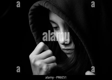 scared girl in hood looking down on black background, monochrome Stock Photo