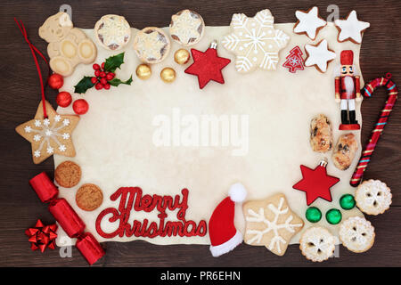 Merry Christmas background border with sign, tree decorations, biscuits, cakes, winter flora  and chocolates in foil on parchment paper on rustic wood Stock Photo