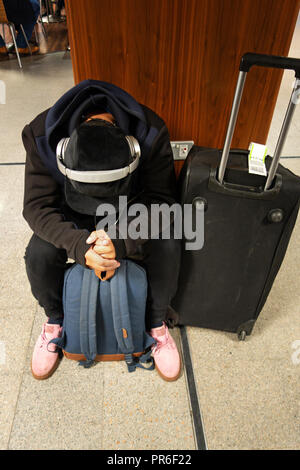 Young man sits on the floor listening to music on headphones in the Arrival Hall at Manchester International Airport, UK Stock Photo